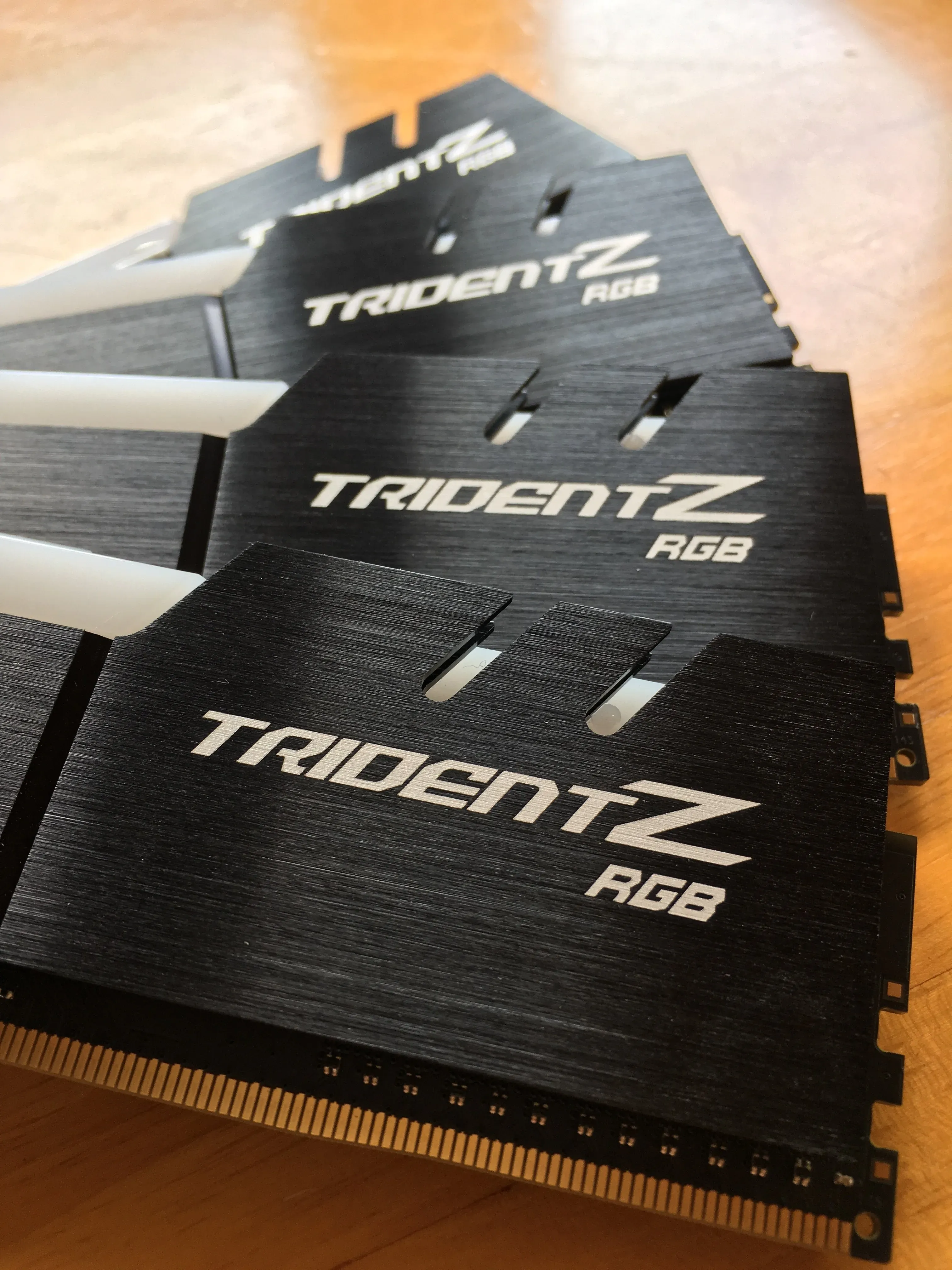 Three Trident Z RGB sticks fanned out on a wooden table, brushed black heatspreaders and gold contacts