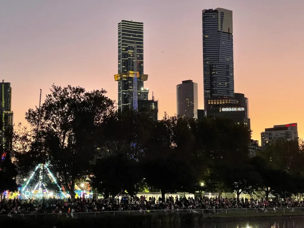 A crowd packed along the riverbank at dusk below lit carnival rides, Southbank towers and the Herald Sun sign beyond