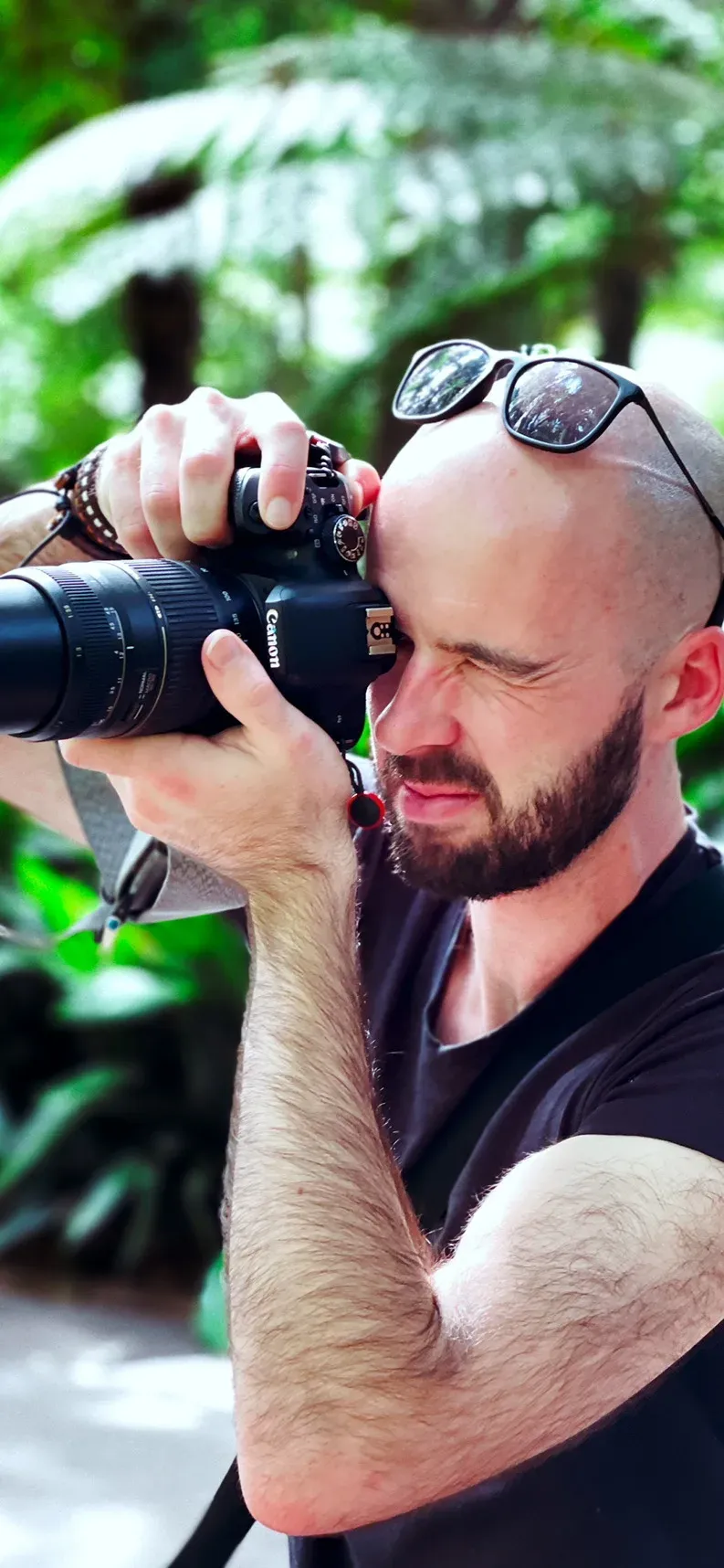 Man with sunglasses pushed up on his head shooting with a Canon DSLR and long lens among tree ferns