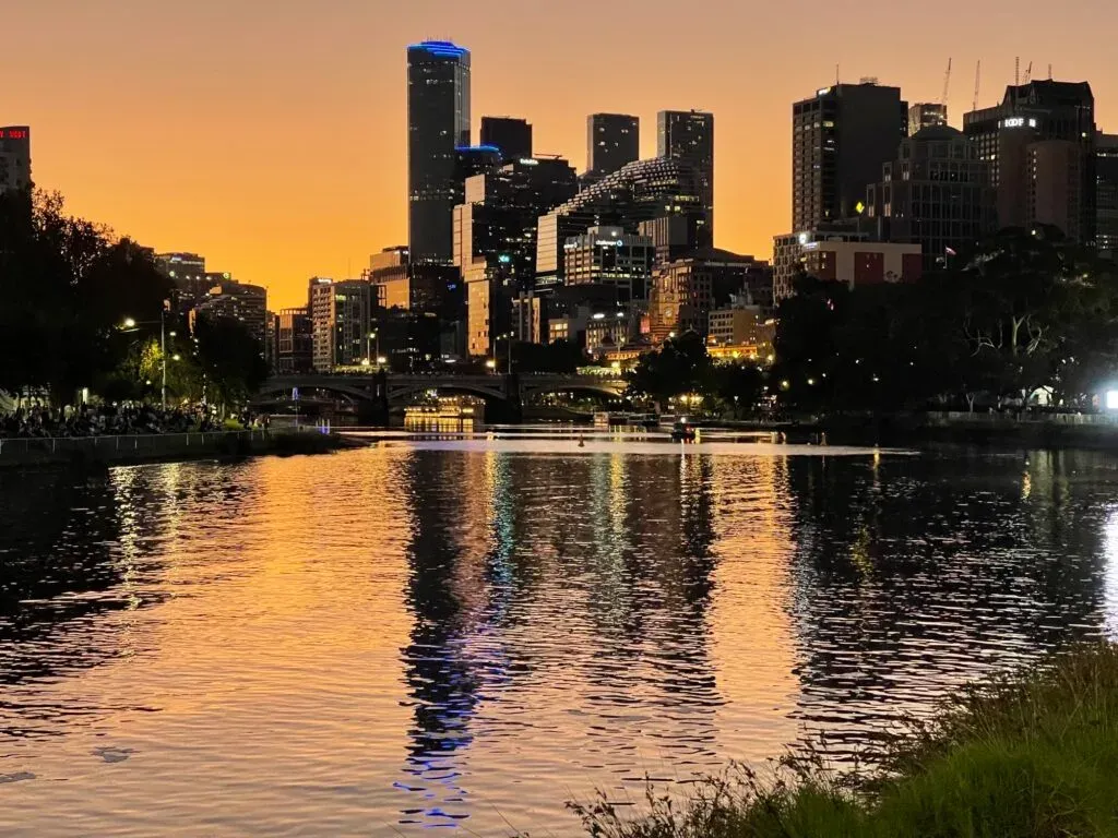 The skyline silhouetted against an orange sunset, city lights streaking down the Yarra, people gathered on the bank