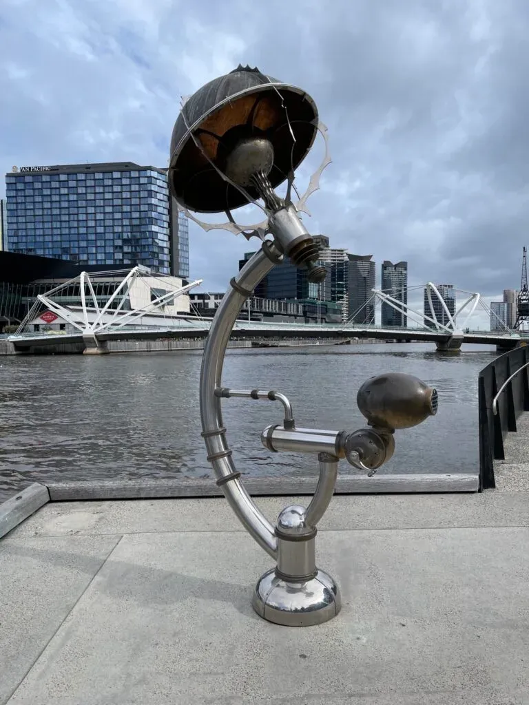 A polished steel sculpture shaped like an oversized lamp on the river promenade, white truss bridge behind
