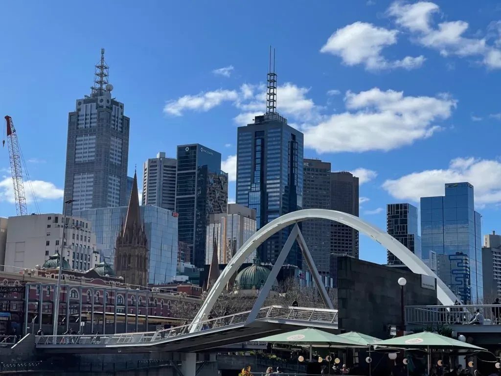 A white arched footbridge over the Yarra below the skyline on a clear blue day, cafe umbrellas beneath