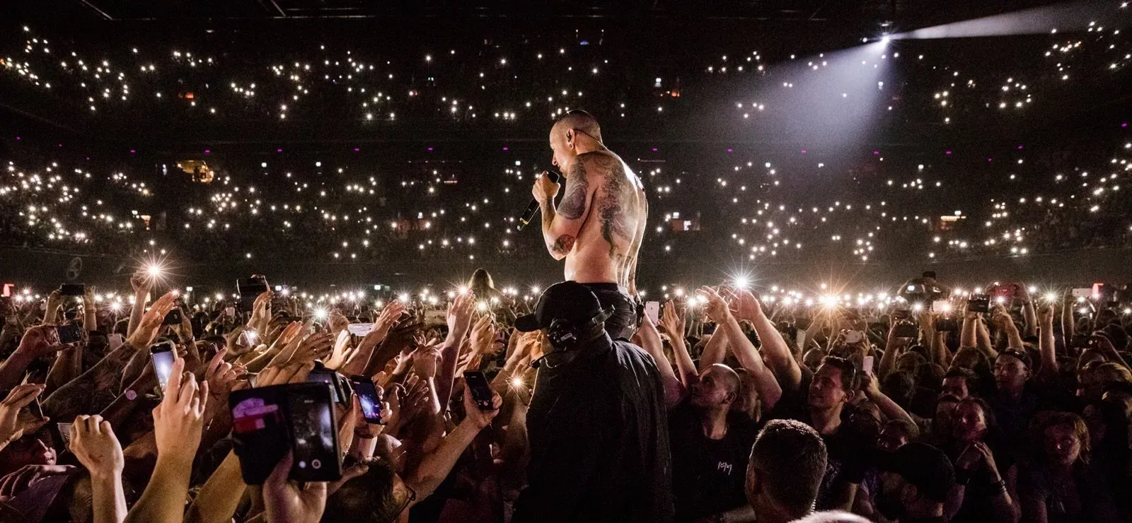 Chester Bennington on stage, shirtless with a microphone, standing above a packed arena floor. Thousands of phone lights fill the crowd and the stands behind him.