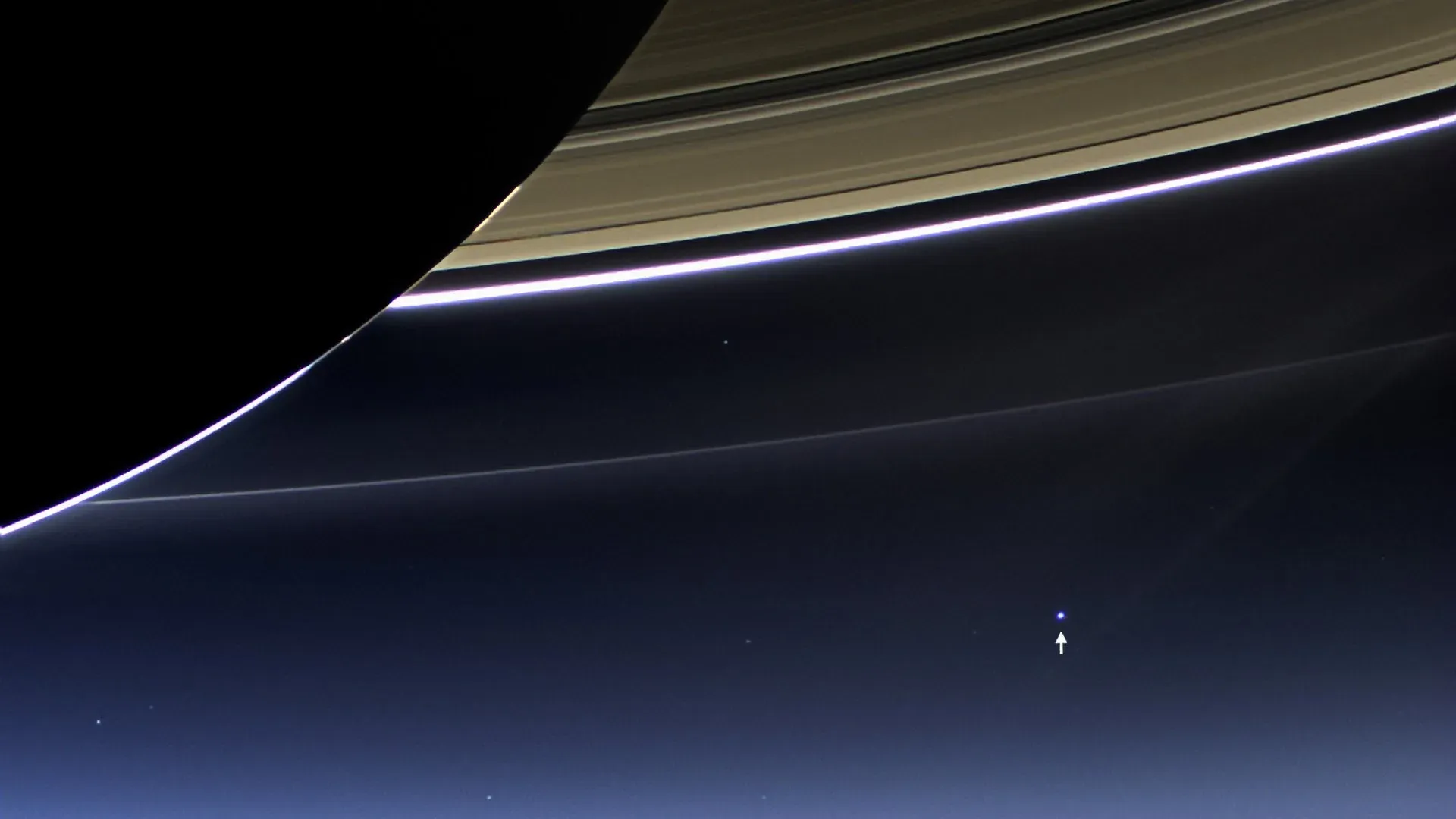 Cassini's view from behind Saturn. The planet's dark limb fills the left, its rings arc bright across the top, and far below in the blue a single pale dot is marked with an arrow. That dot is Earth.