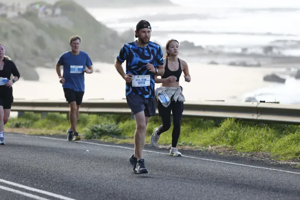 Kyle running the coast road, bib 10059 marked Kyle Team, surf breaking over rocks behind him