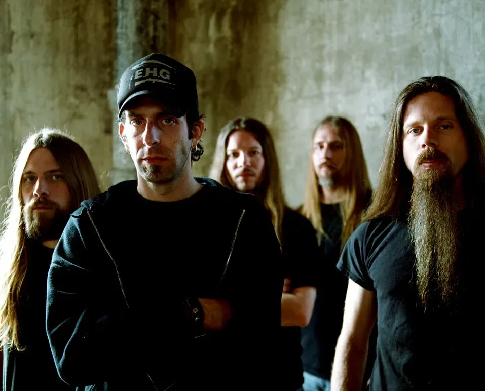 The five members of Lamb of God standing against a concrete wall, mostly long hair and black shirts, the singer in front in a cap with his arms folded.