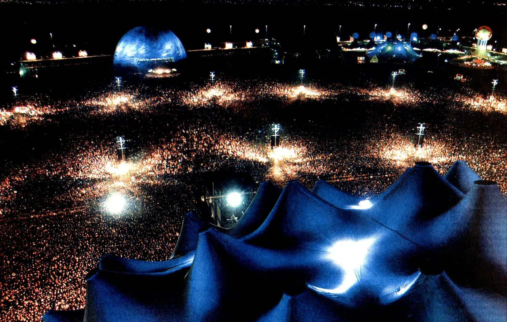 Night aerial view over a festival field packed with a crowd to the horizon, floodlight towers and blue tent roofs