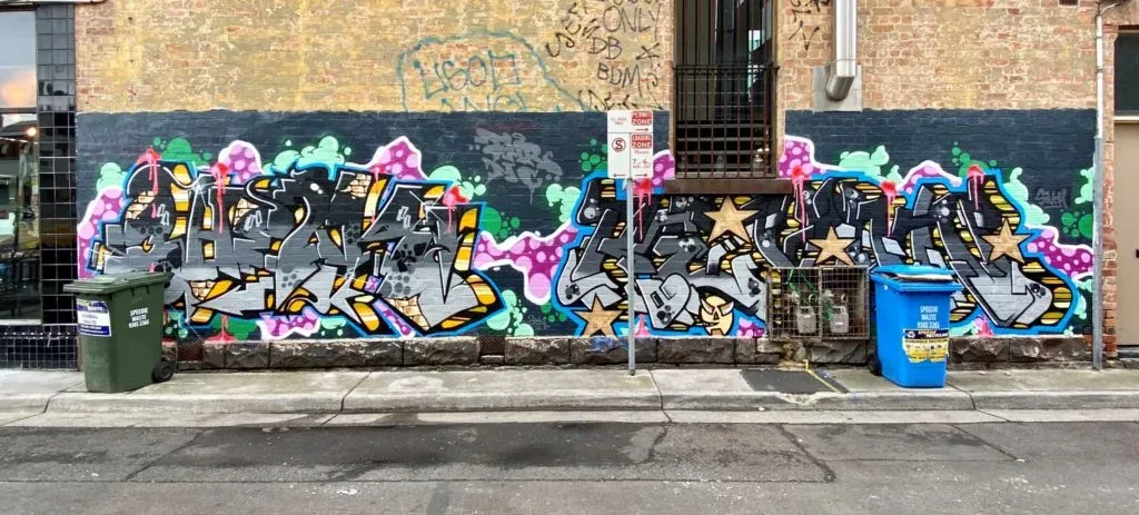 Wide graffiti piece in chunky silver letters with gold stars, painted across a brick laneway wall between two wheelie bins