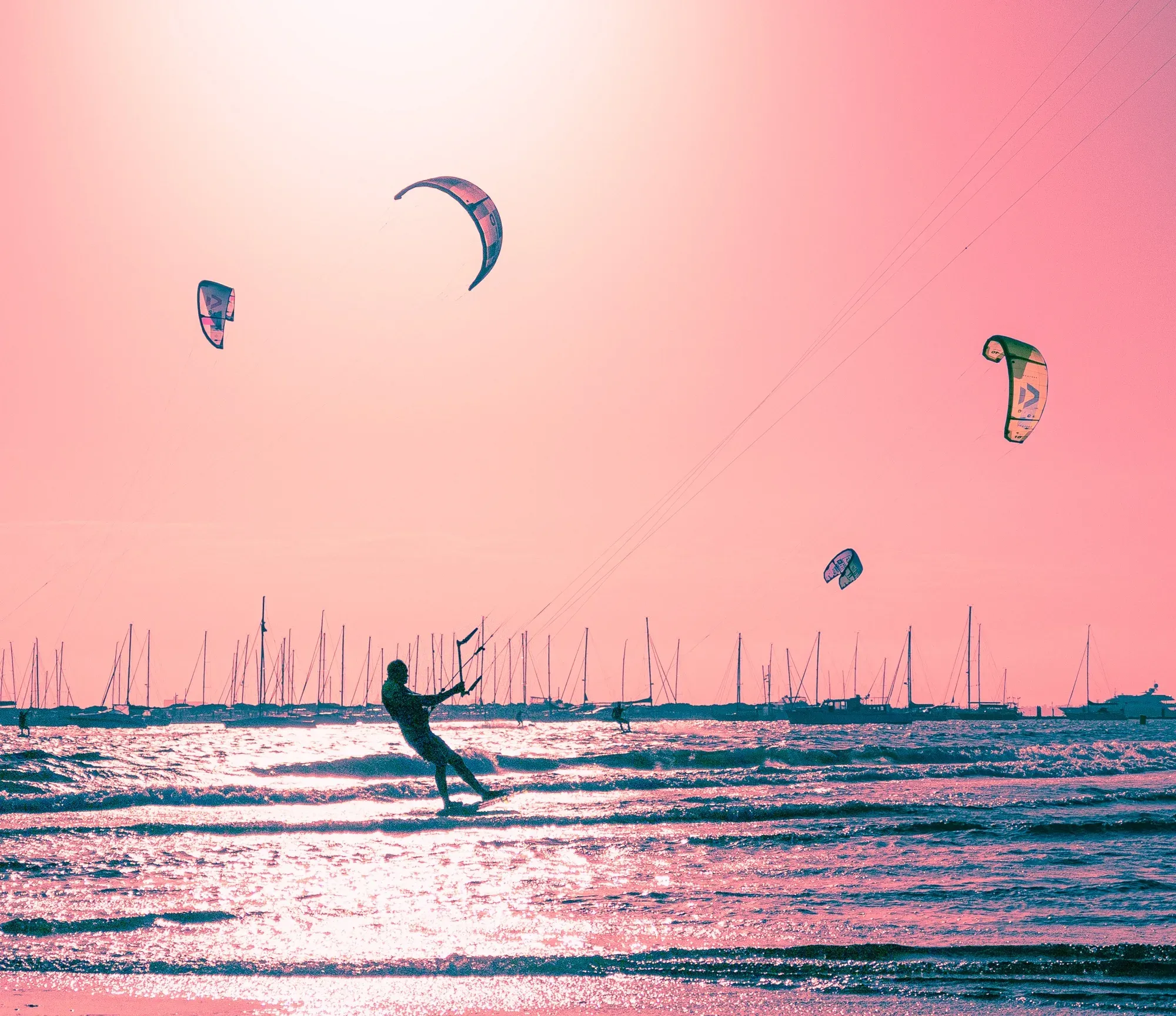 Kitesurfer cutting across the shallows past a forest of moored yacht masts, the sky washed pink