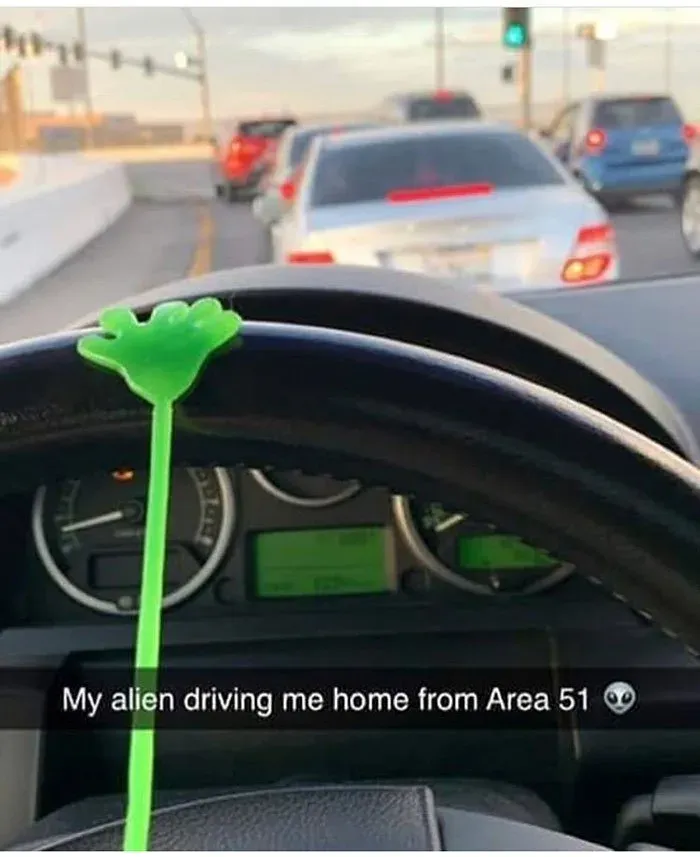 Green plastic alien hand stuck to a car steering wheel in traffic, captioned "My alien driving me home from Area 51"