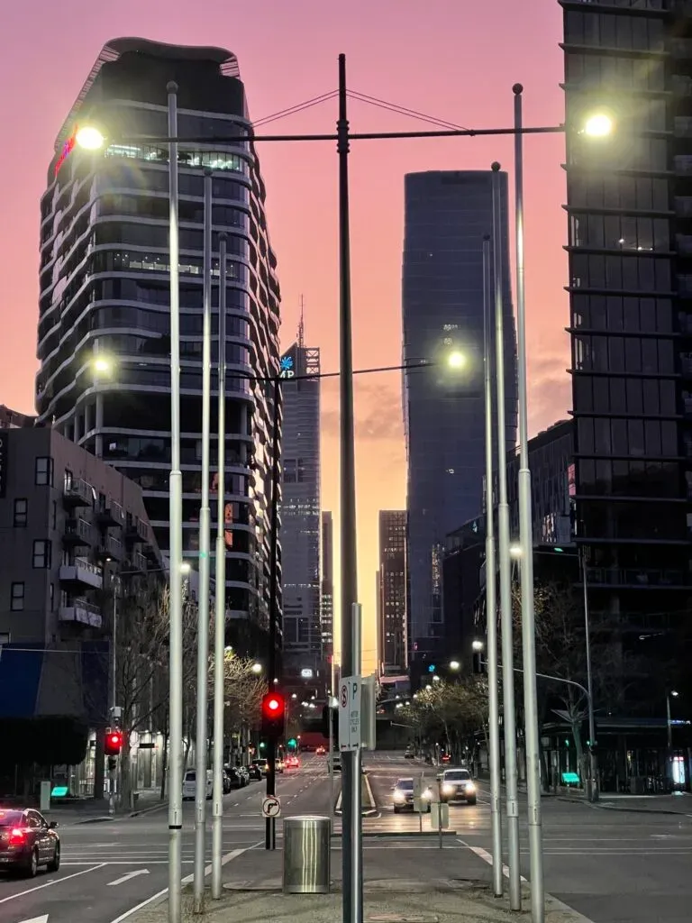 A near-empty city street at dawn, streetlights still burning against a pink and orange sky between towers