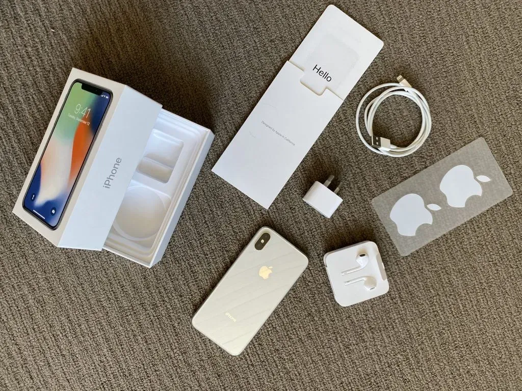 Silver iPhone X face down on carpet beside its open box, EarPods, cable, charger, Hello leaflet and Apple stickers