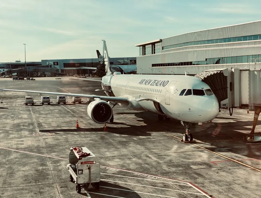 Air New Zealand jet parked at an airport gate with the airbridge attached and baggage carts alongside