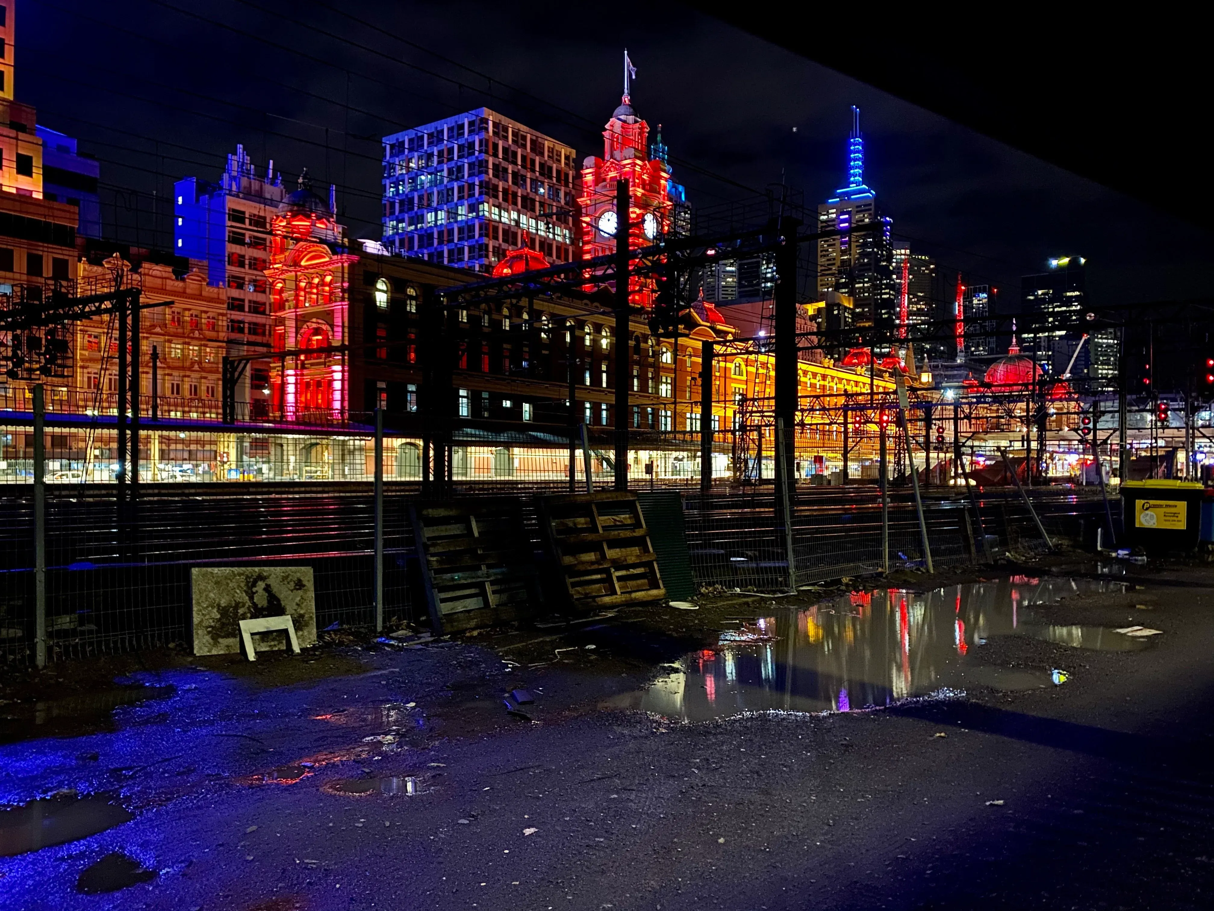 Flinders Street Station lit red and gold at night, seen across the rail yard with the city skyline behind