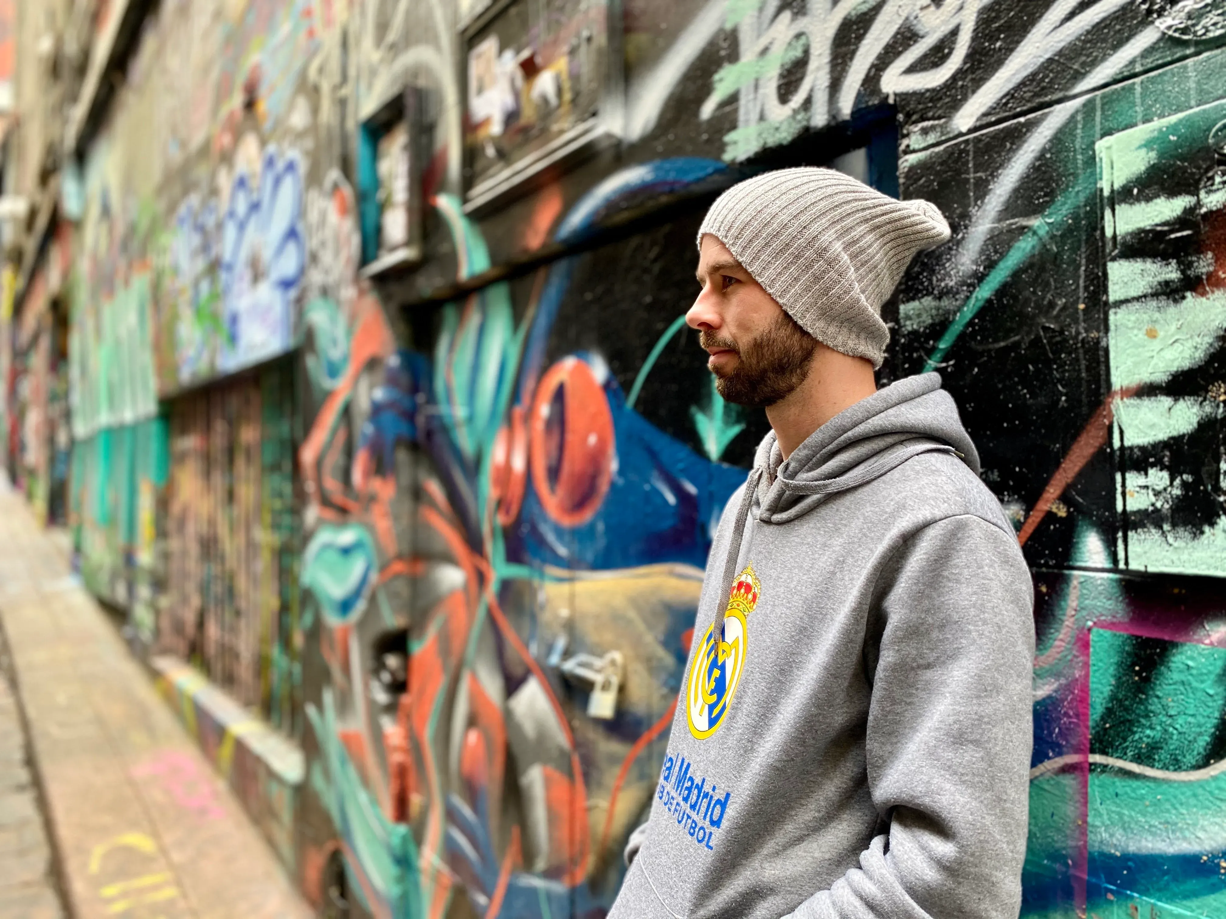 Jeremy in a grey beanie and Real Madrid hoodie, looking down a laneway covered wall to wall in graffiti