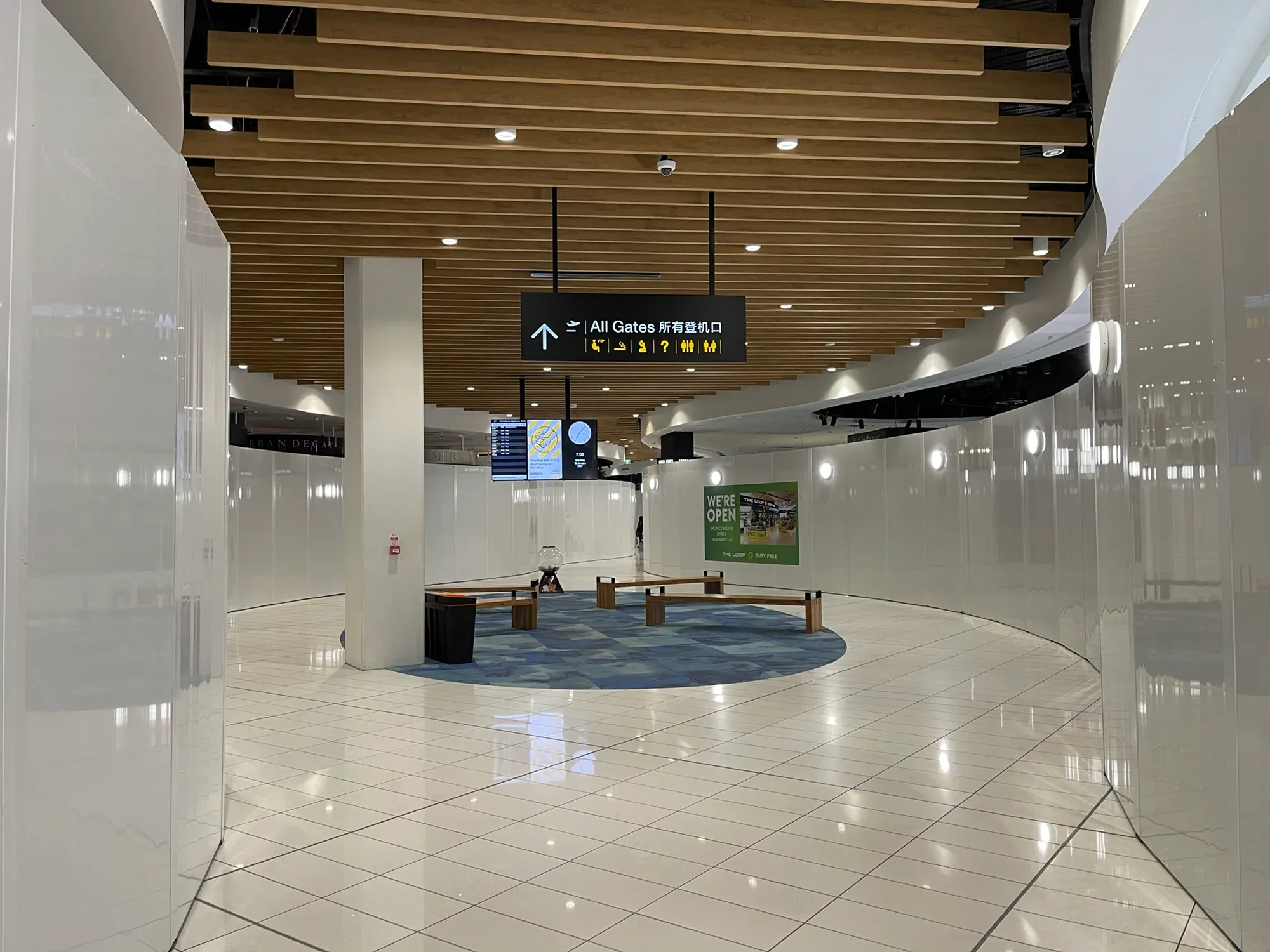 An empty white-panelled walkway under an All Gates sign, one "We're open" duty free poster left