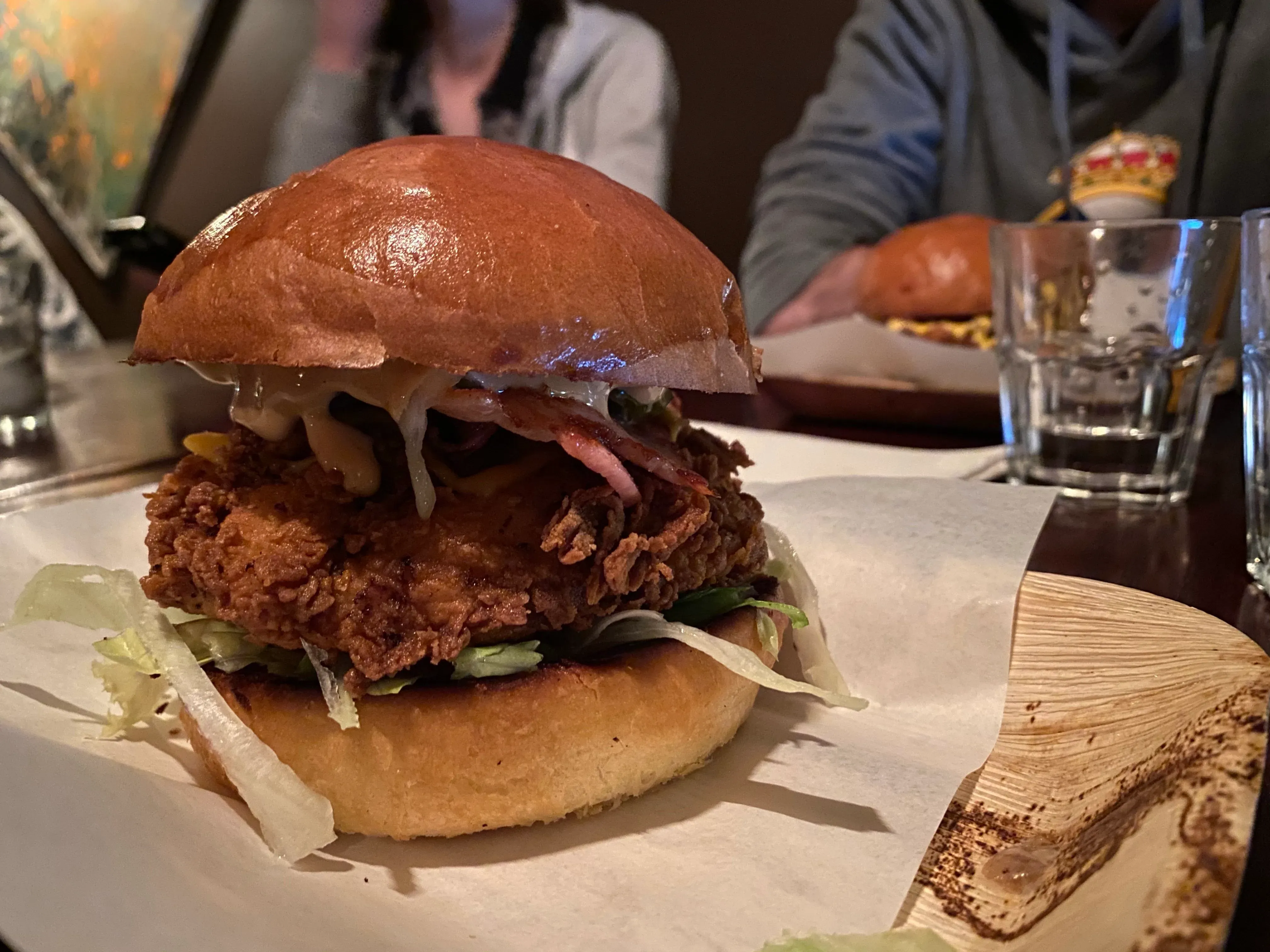 Crumbed chicken fillet with melted cheese, bacon and lettuce in a glossy brioche bun, served on paper at a dim table