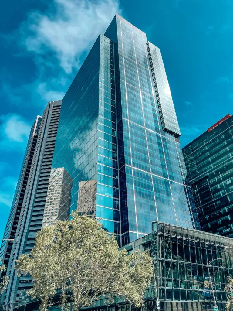 Looking up the mirrored blue face of a glass tower, an Australia Post sign on the block beside it