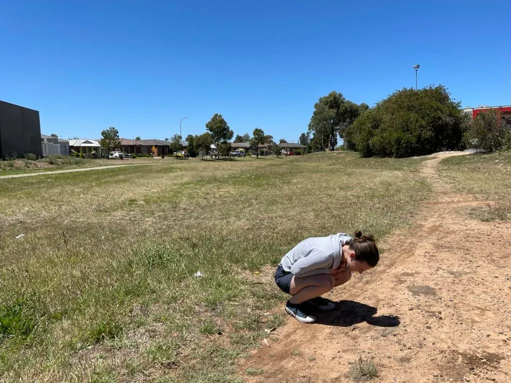 Simone crouched low on a dirt track through dry grass, houses at the reserve's edge