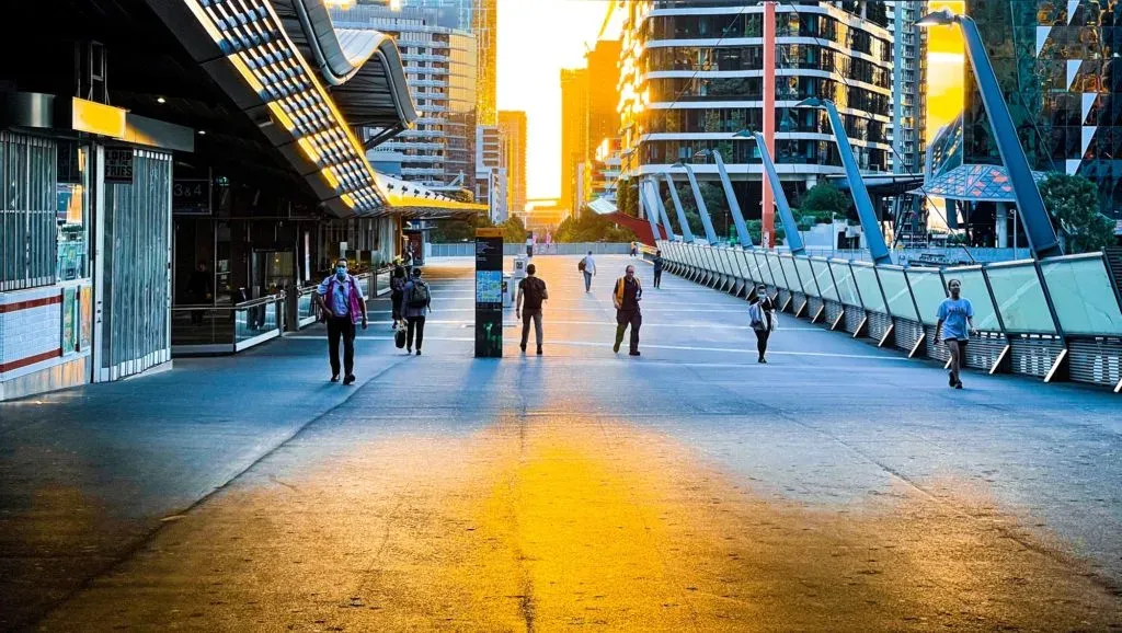 Masked commuters crossing a wide pedestrian bridge into a golden sunset between glass towers