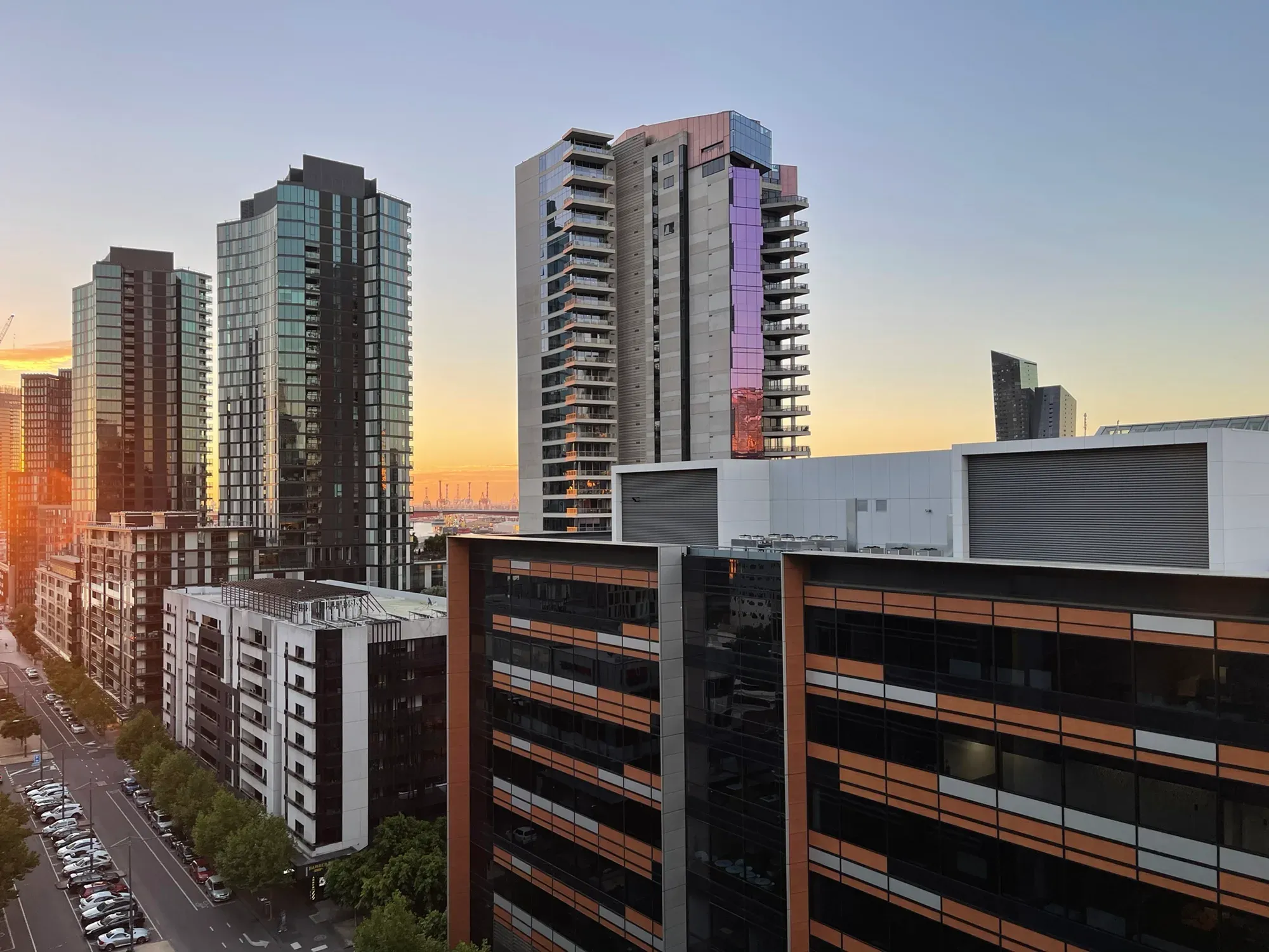 Docklands apartment towers at sunset, a street of parked cars below and port cranes on the horizon