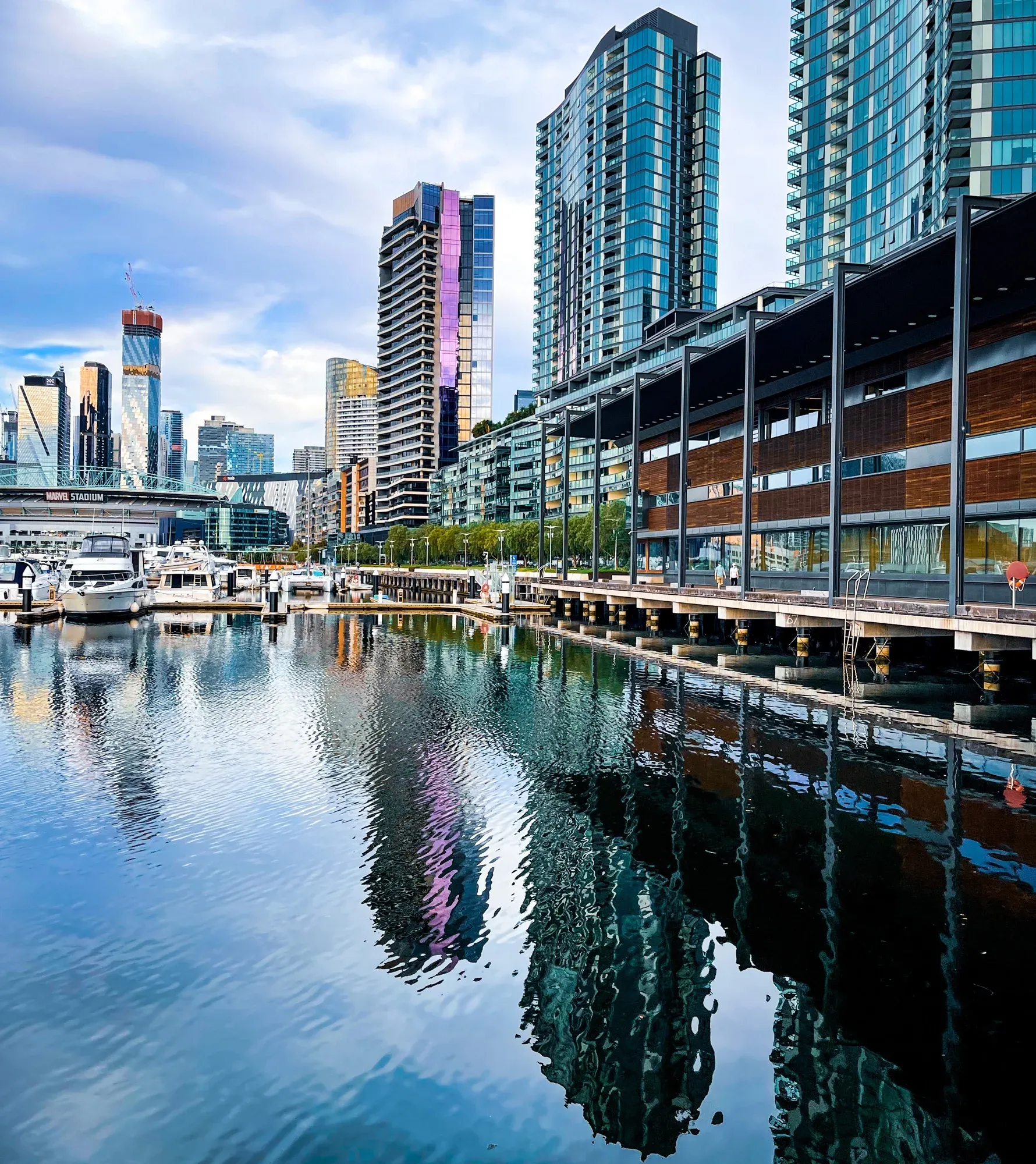 Marvel Stadium and the Docklands towers mirrored in flat marina water, boats moored at the far dock