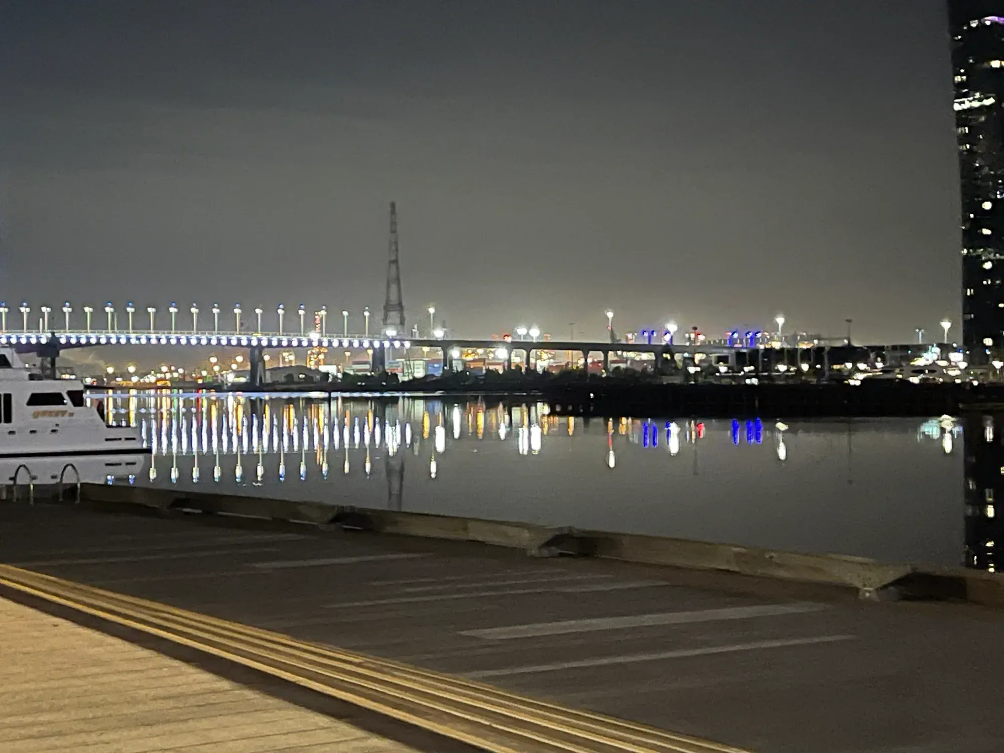 Lamp-lit bridge across the harbour at night, its lights mirrored in flat water beside a moored cruiser