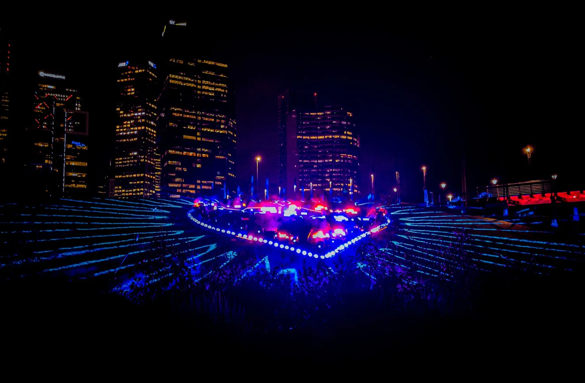 Heart-shaped light sculpture glowing blue and pink on a terraced lawn below the office towers