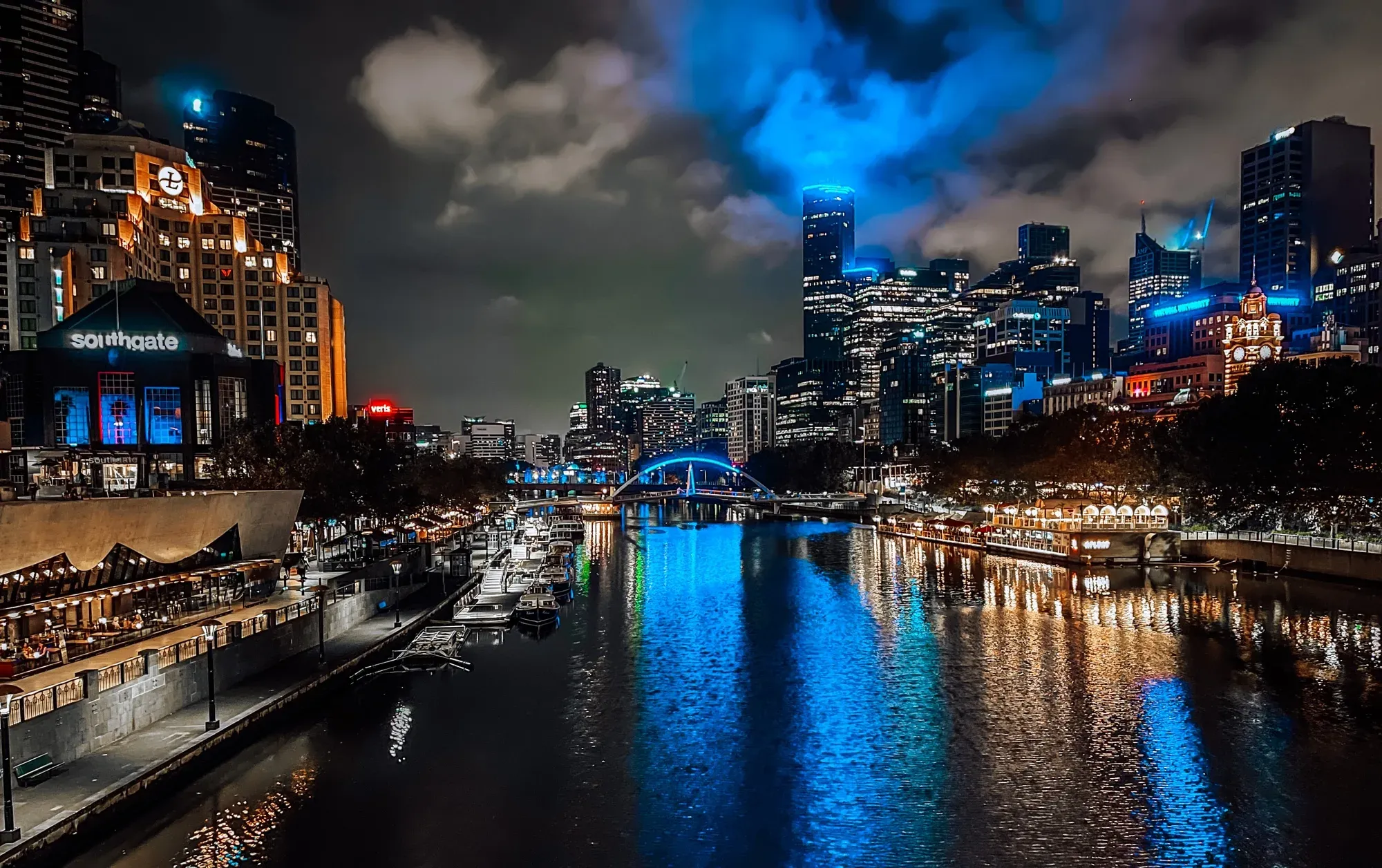 Southgate and the CBD towers lit at night above the Yarra, an arched bridge glowing blue upstream