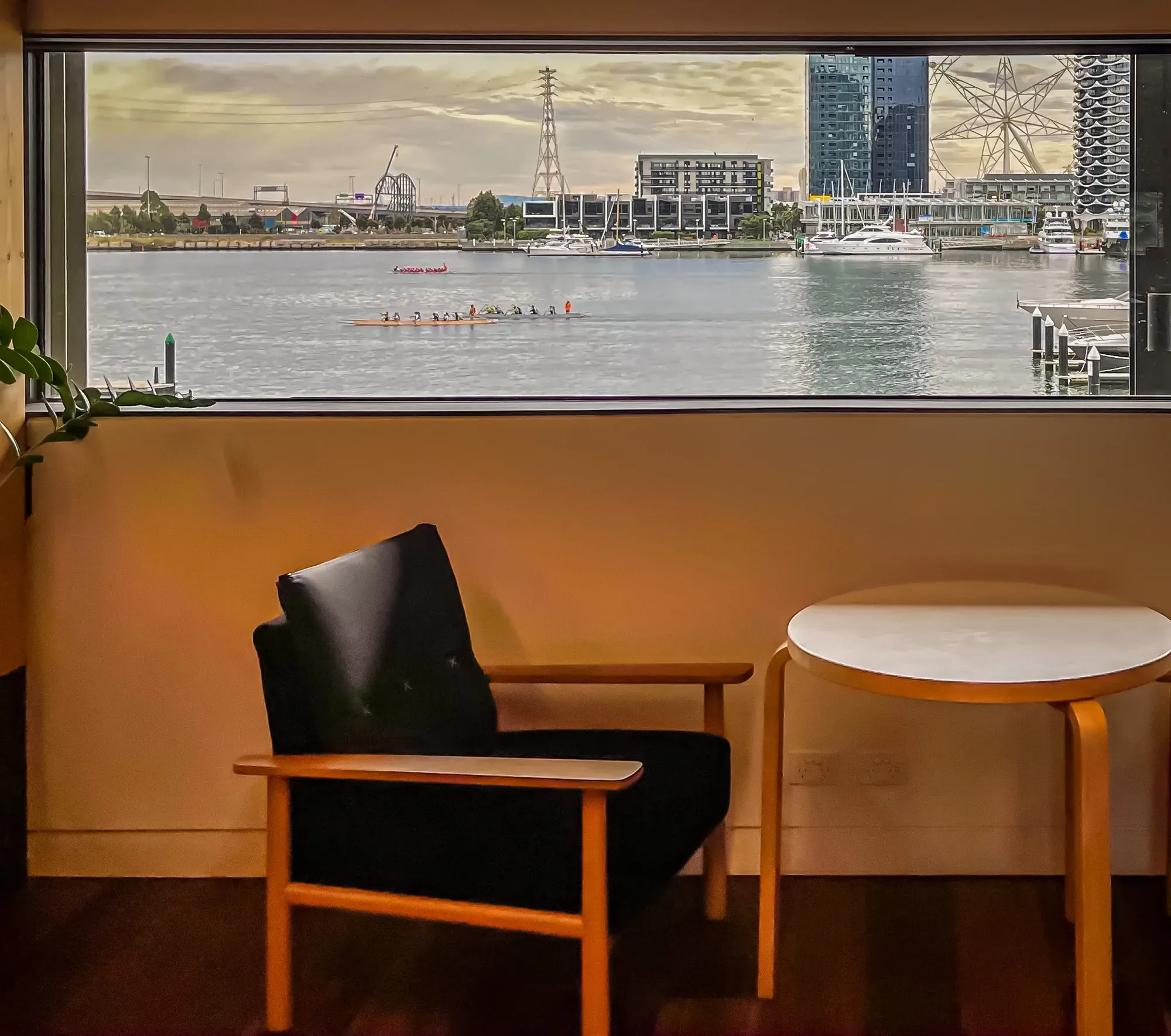 Rowing crews and a dragon boat on the harbour, framed by a wide window above an empty armchair and side table