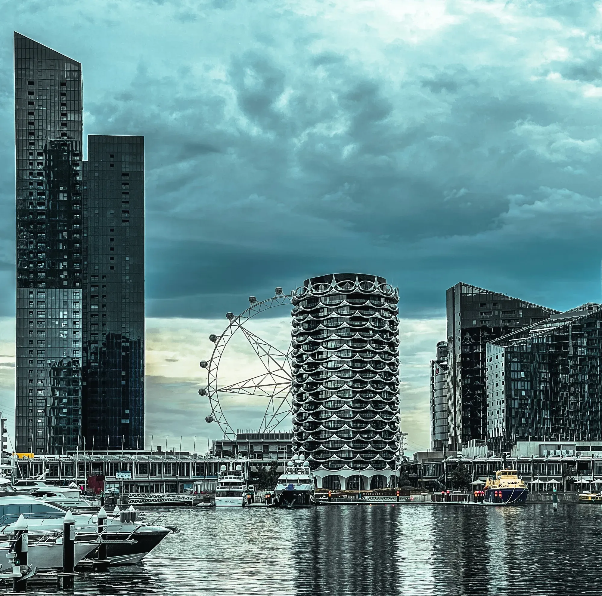Docklands observation wheel and a scalloped white apartment tower above moored boats, under heavy cloud