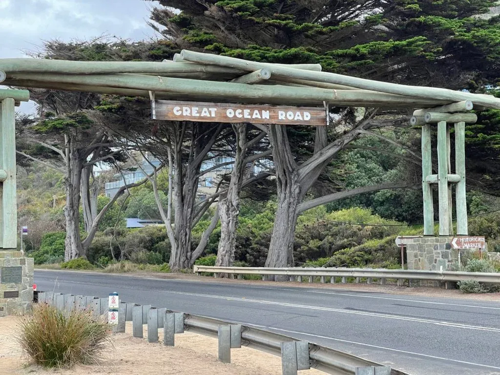 Rough timber arch over the roadside, its painted sign reading Great Ocean Road, wind-bent cypress behind