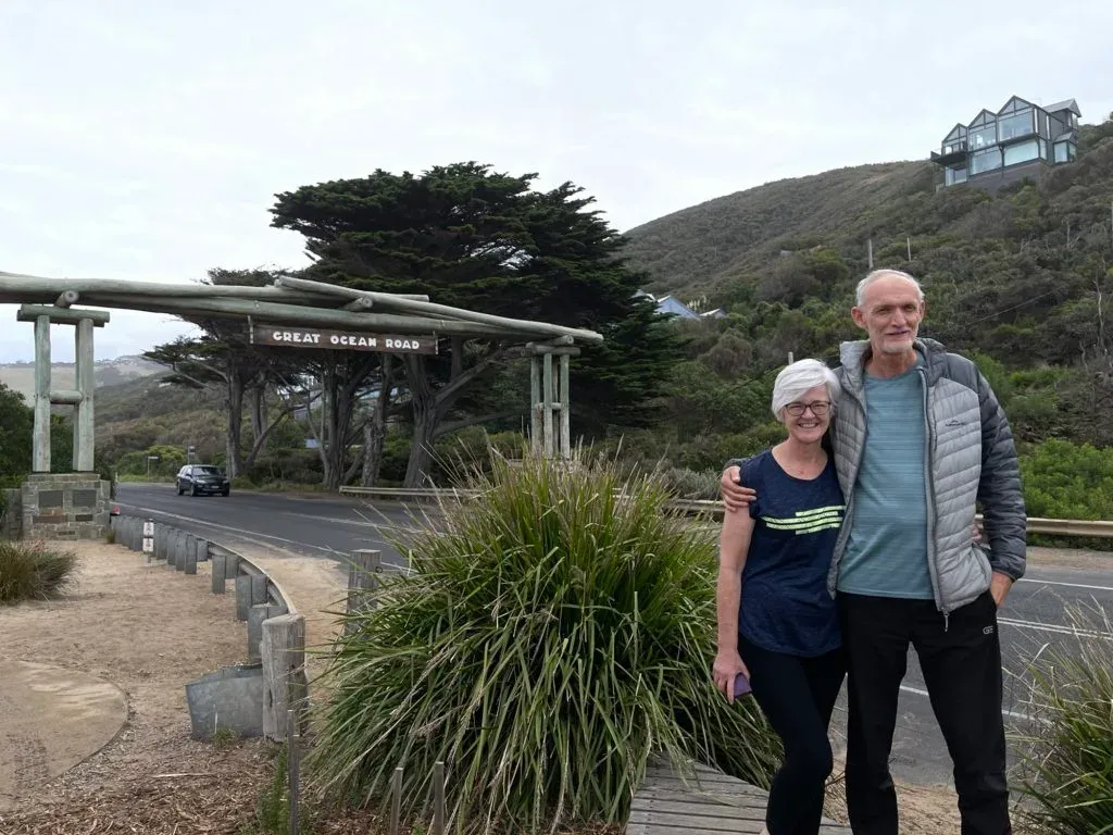 An older couple standing arm in arm by the roadside, the timber arch and a green headland behind them