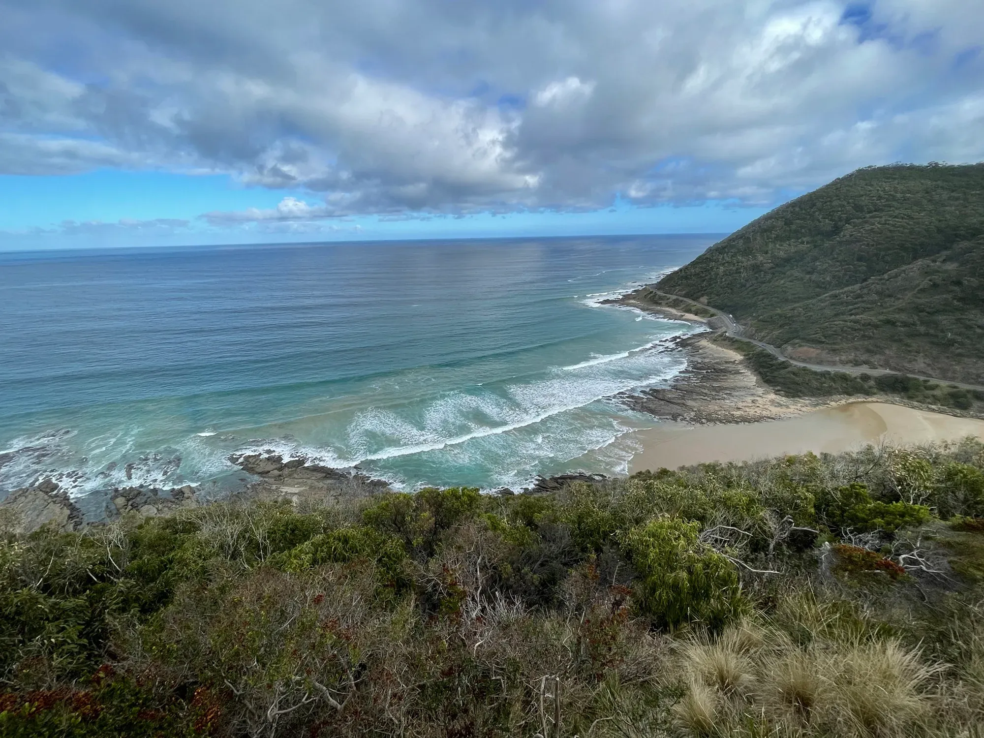 Surf rolling onto a sandy beach far below, a scrub-covered headland with the road cut into its side