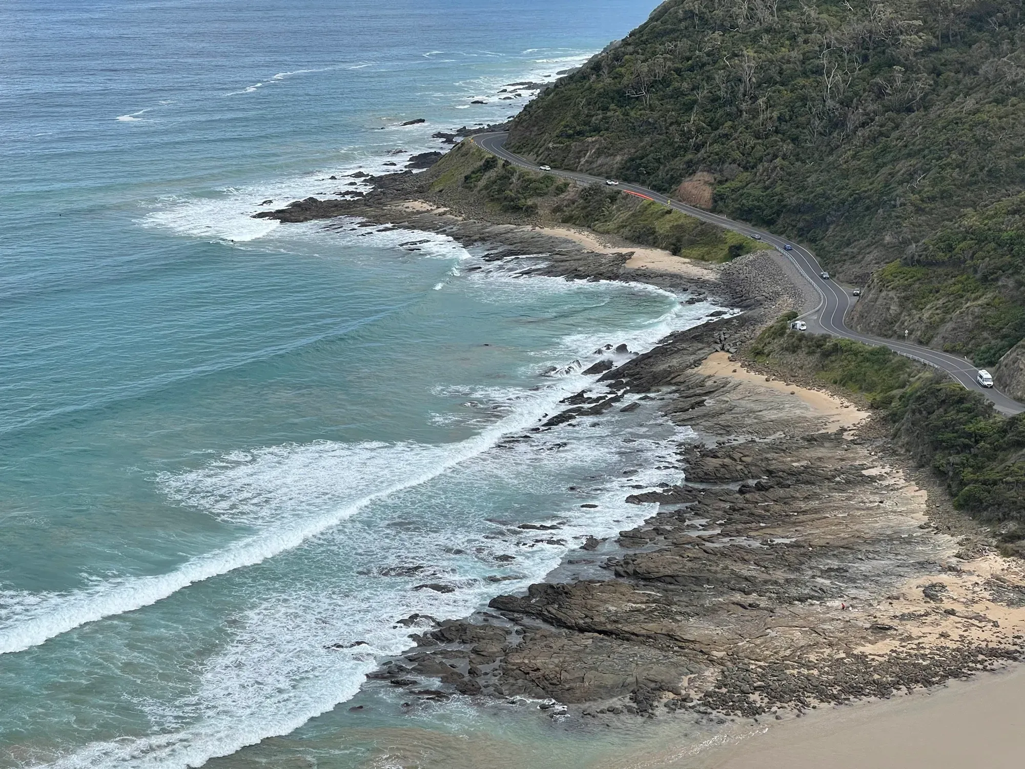 White surf breaking over rock shelves below, the road threading along the hillside above the sand