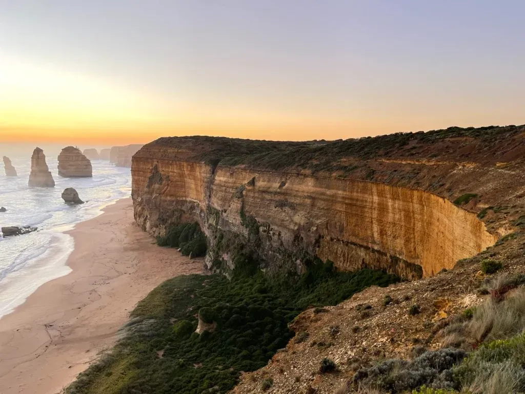 A tall layered cliff face lit gold along the beach, more stacks in the haze beyond