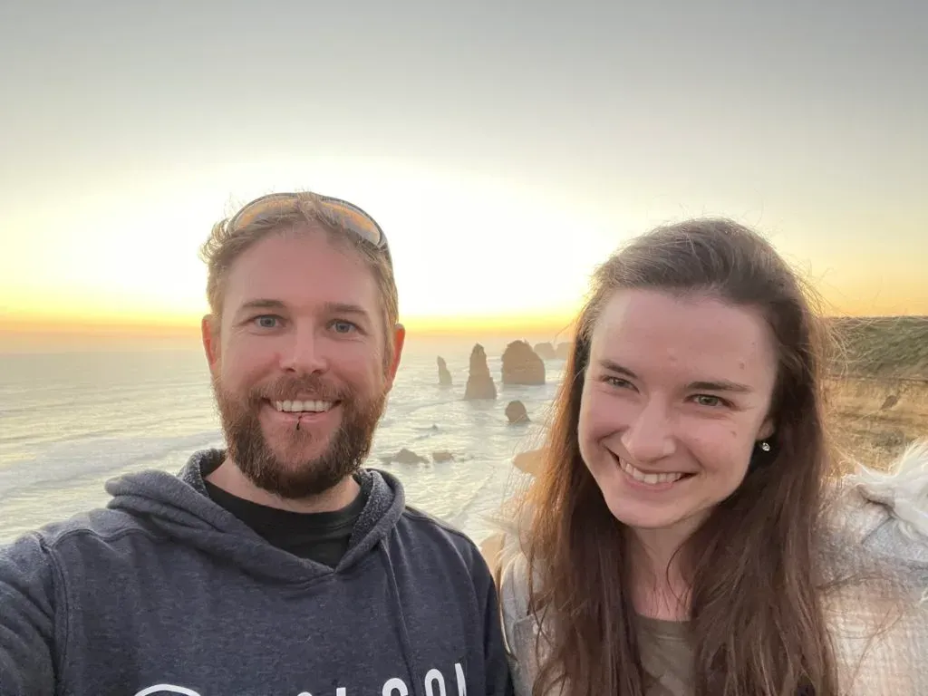 Two people smiling close to the camera, the stacks and a gold sunset over the water behind them