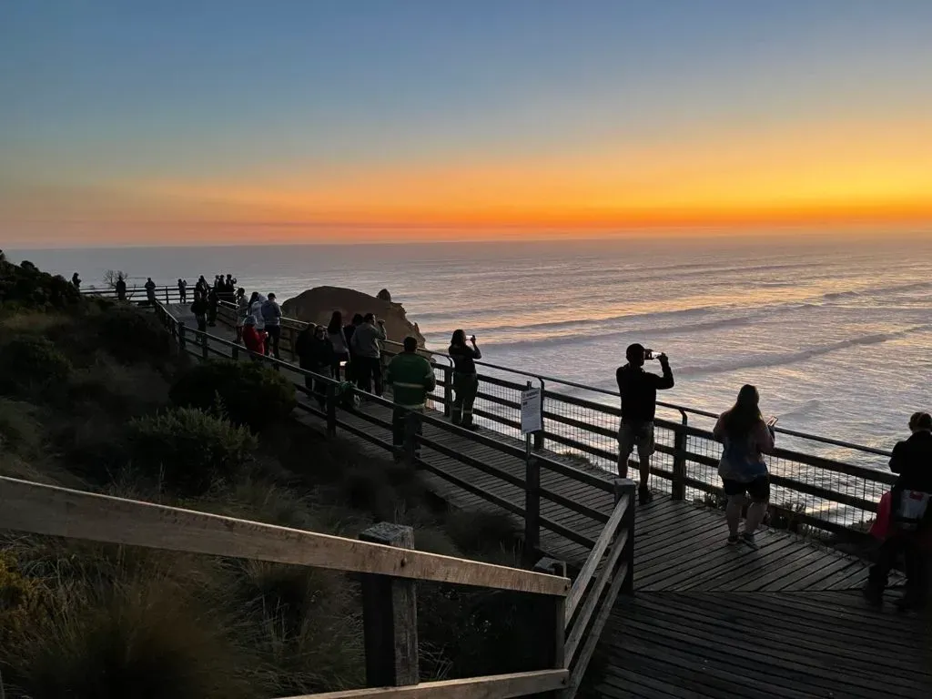 Visitors lined along the boardwalk railing photographing the sunset over the ocean