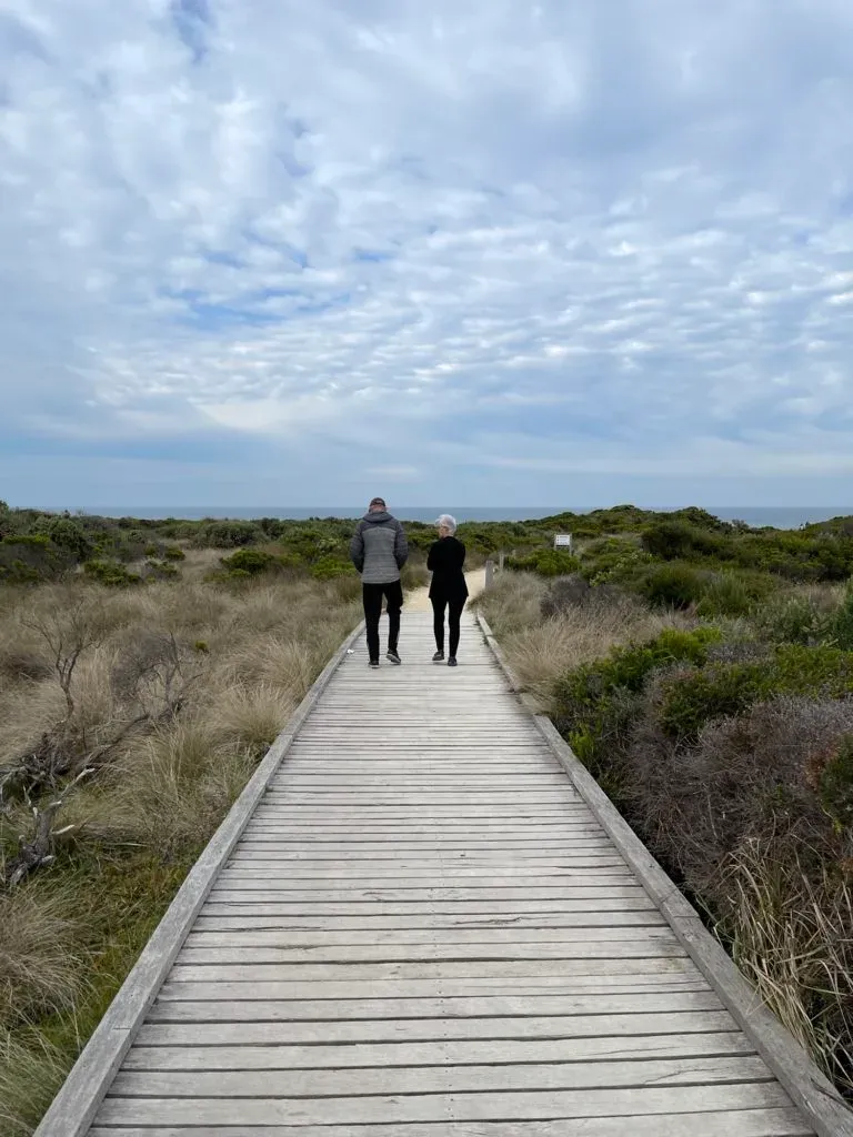 A couple walking a long timber boardwalk through low coastal scrub towards the sea