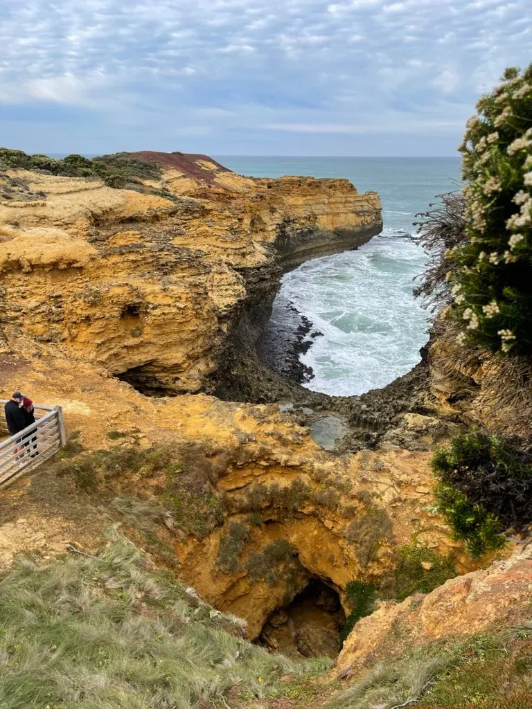 Two people at a timber rail above a collapsed limestone cove, surf pushing into the gap