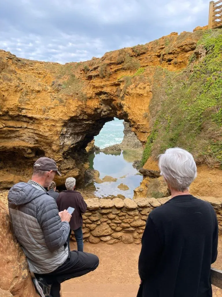 Three visitors at the low stone wall at the bottom of the grotto, one holding a phone up to the arch