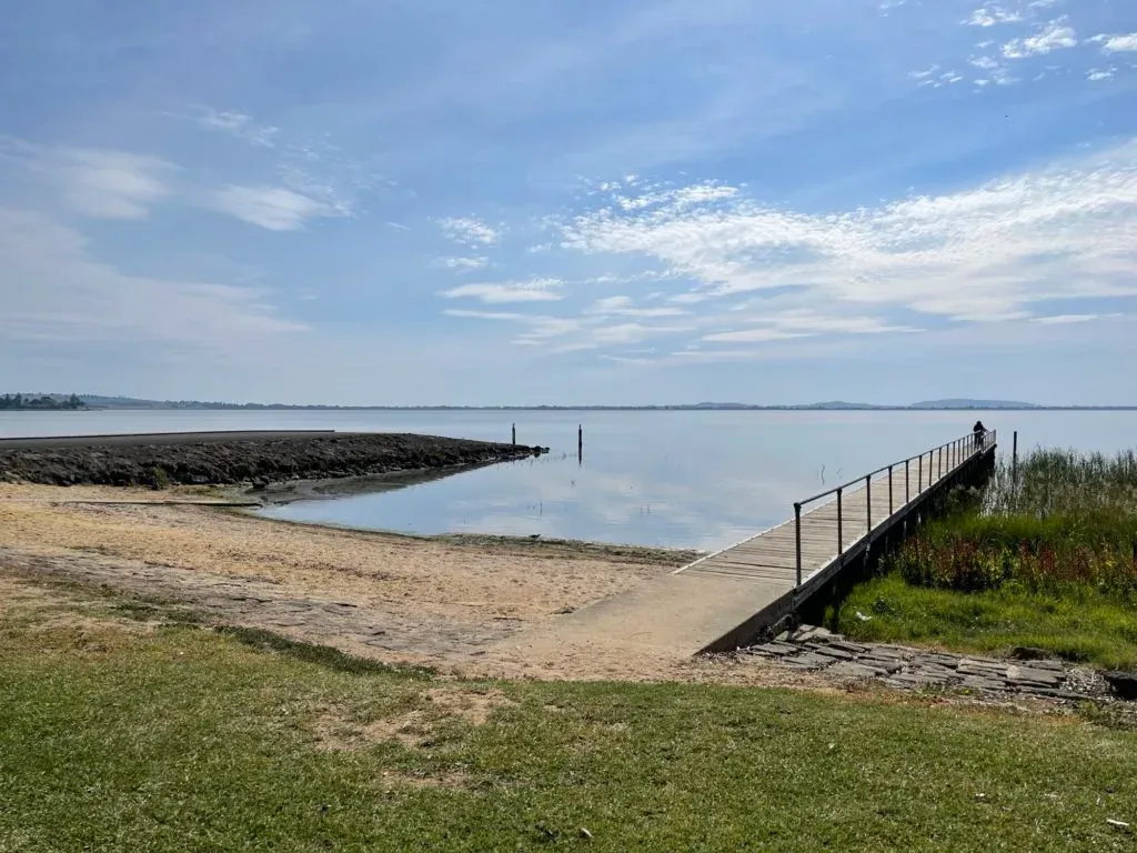 A long jetty reaching out over the flat calm of Lake Colac, sandy shore in front
