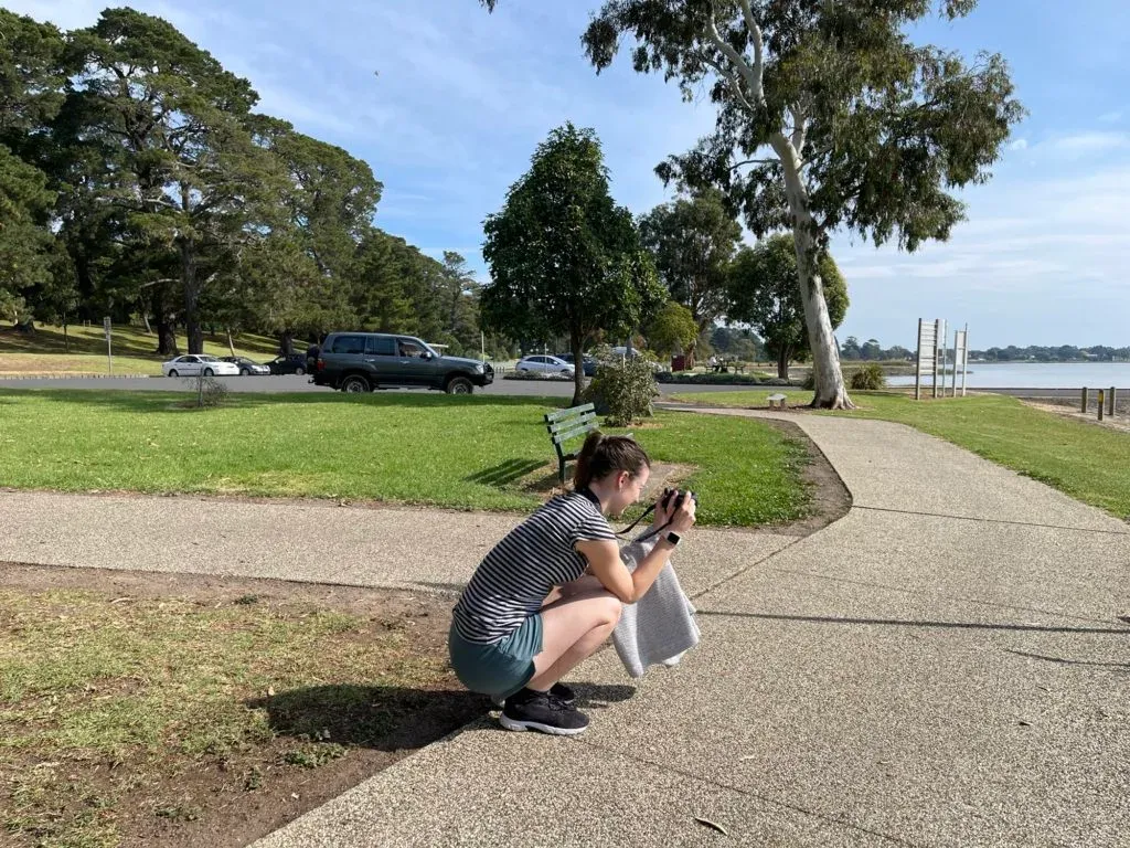 A young woman crouched on a lakeside path with a camera to her eye, gum trees behind her