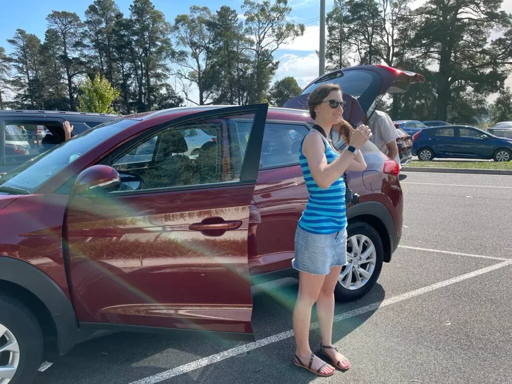 Woman in a striped singlet standing by the open door of a maroon SUV in a sunny car park