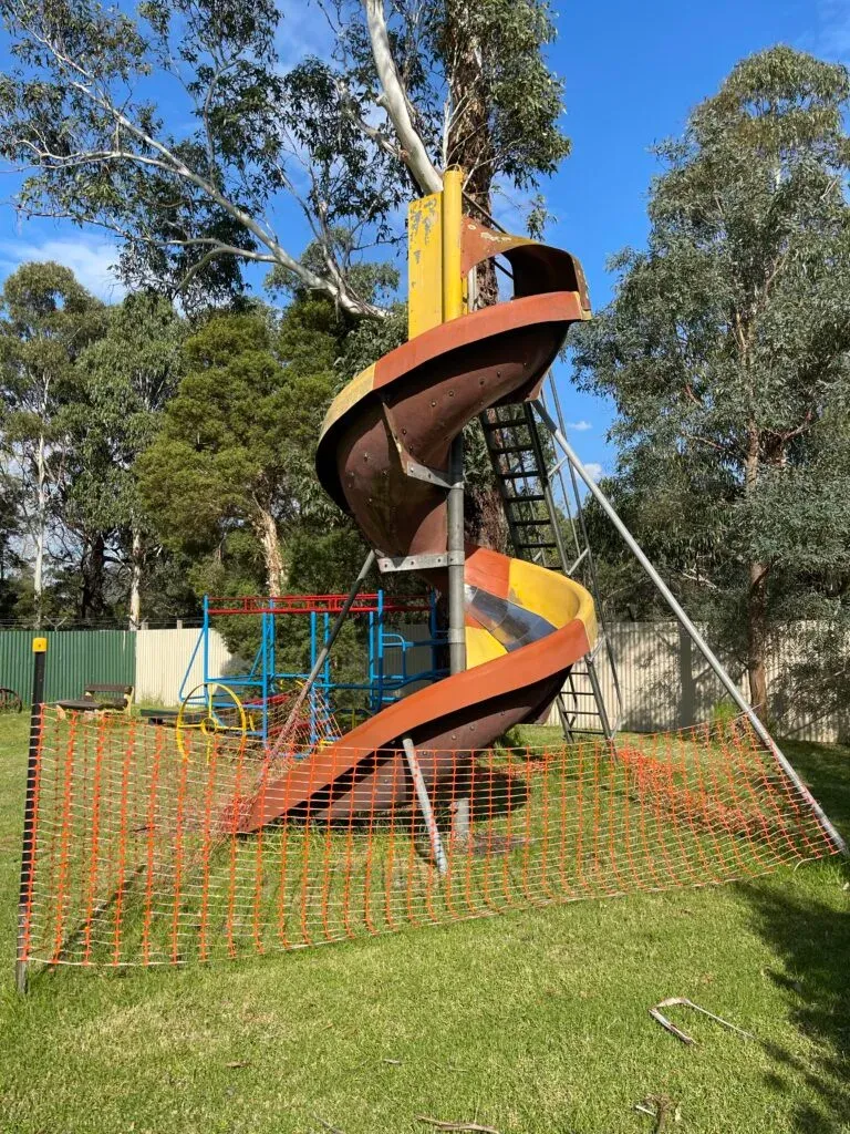 Rusting spiral tube slide fenced off behind orange safety mesh, older playground frames beside it under gums