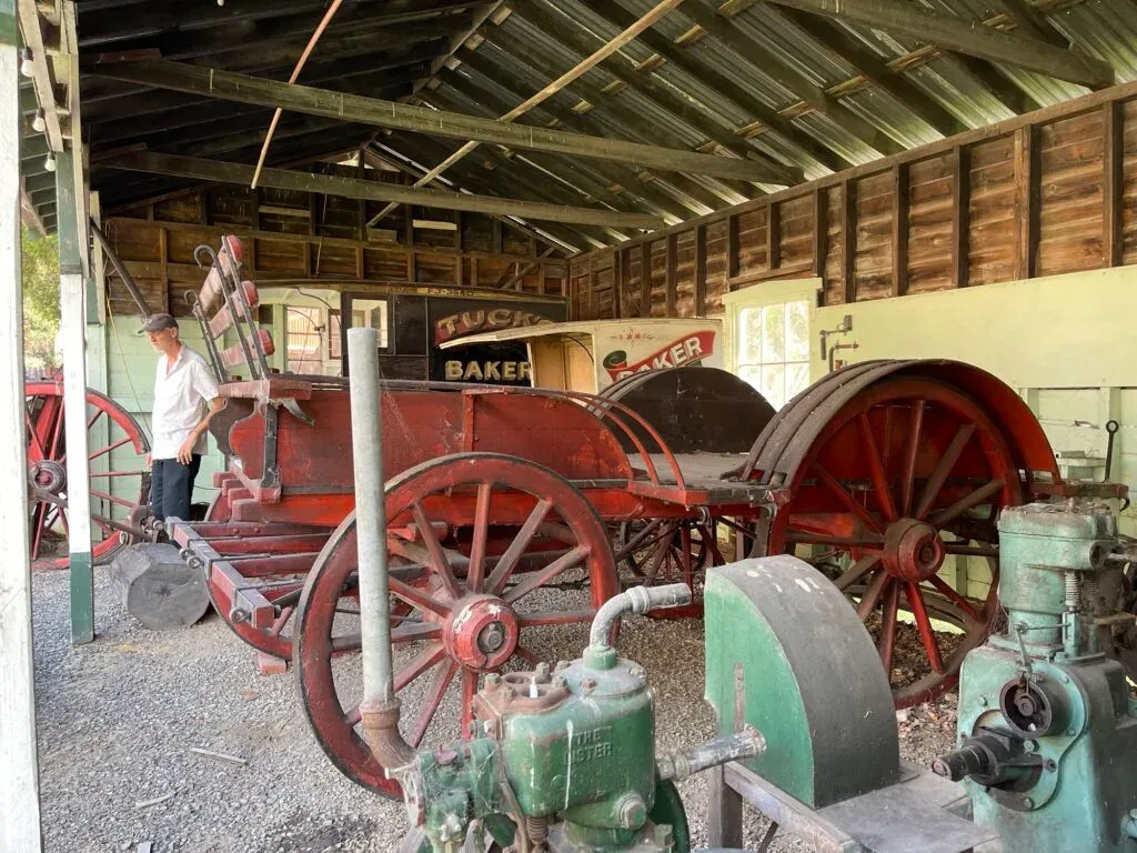 Red-wheeled horse-drawn dray in an open shed, baker's signage behind it and green stationary engines in front