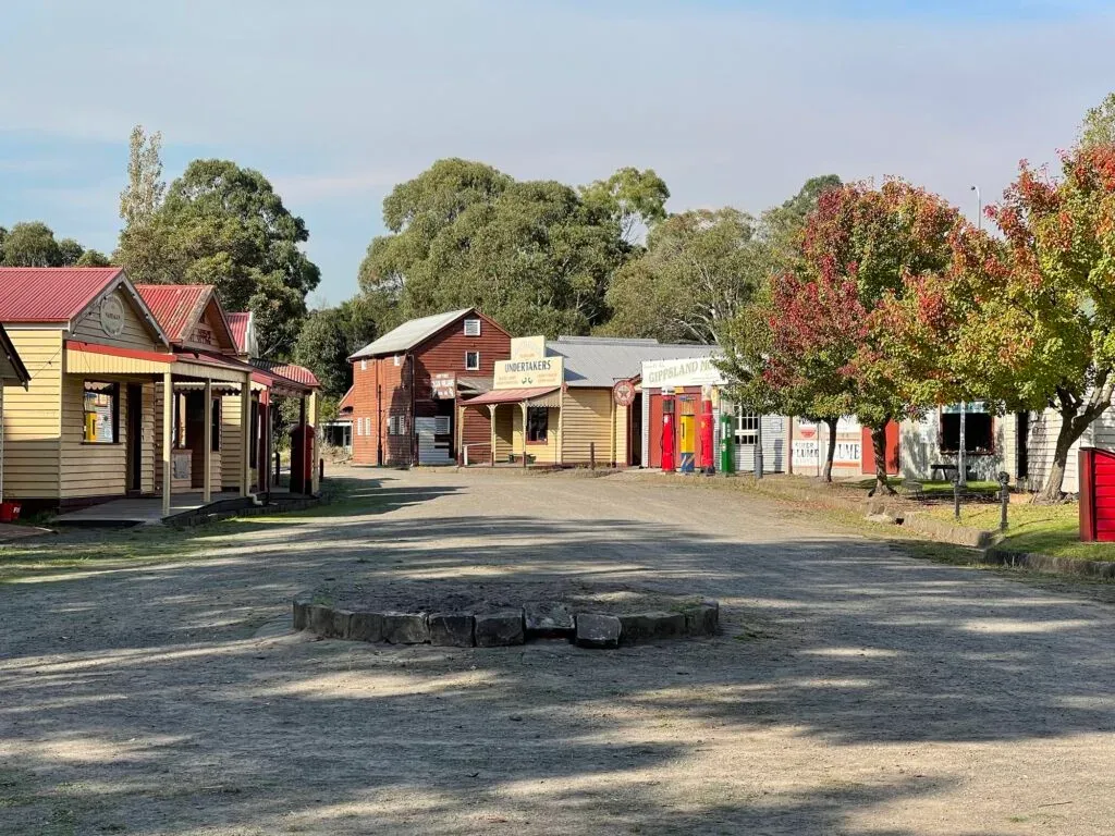 Dirt main street lined with weatherboard shopfronts, an undertaker's among them, a stone fire ring in the foreground