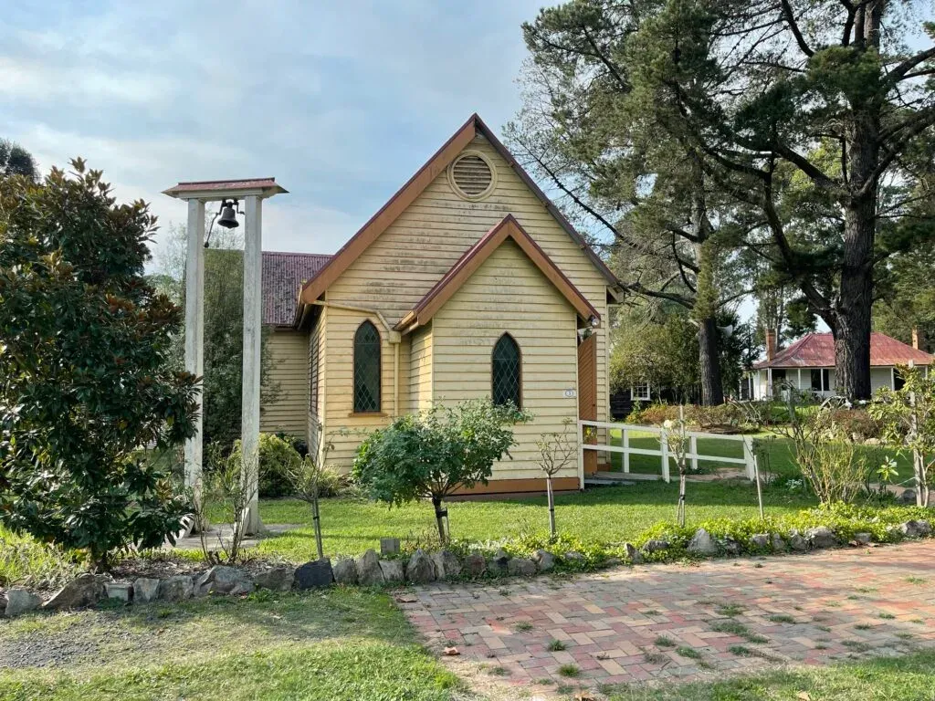 Small yellow weatherboard church with pointed-arch windows and a bell hung in a freestanding timber frame
