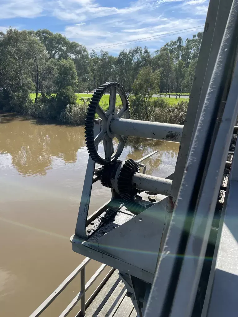Close on the big toothed cog and its pinion in the turning gear, brown river below
