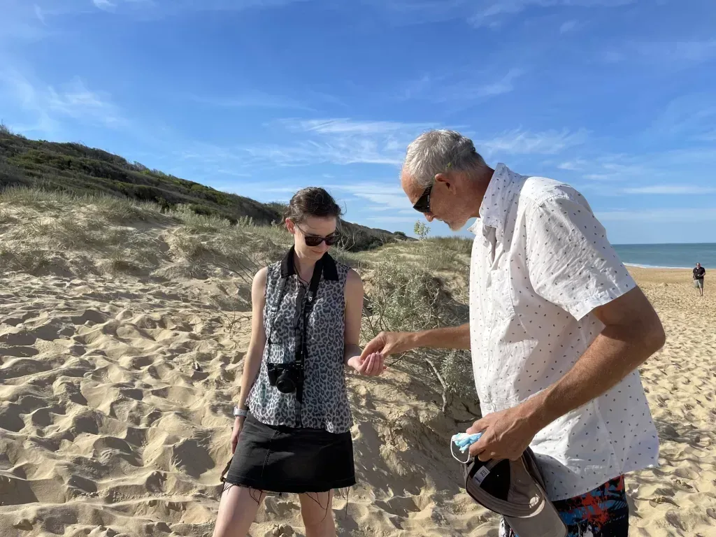 A woman with a camera round her neck peering at something an older man holds out, up on the dunes